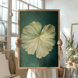 A person is holding up a framed painting of a large, vibrant green leaf against a dark green background.