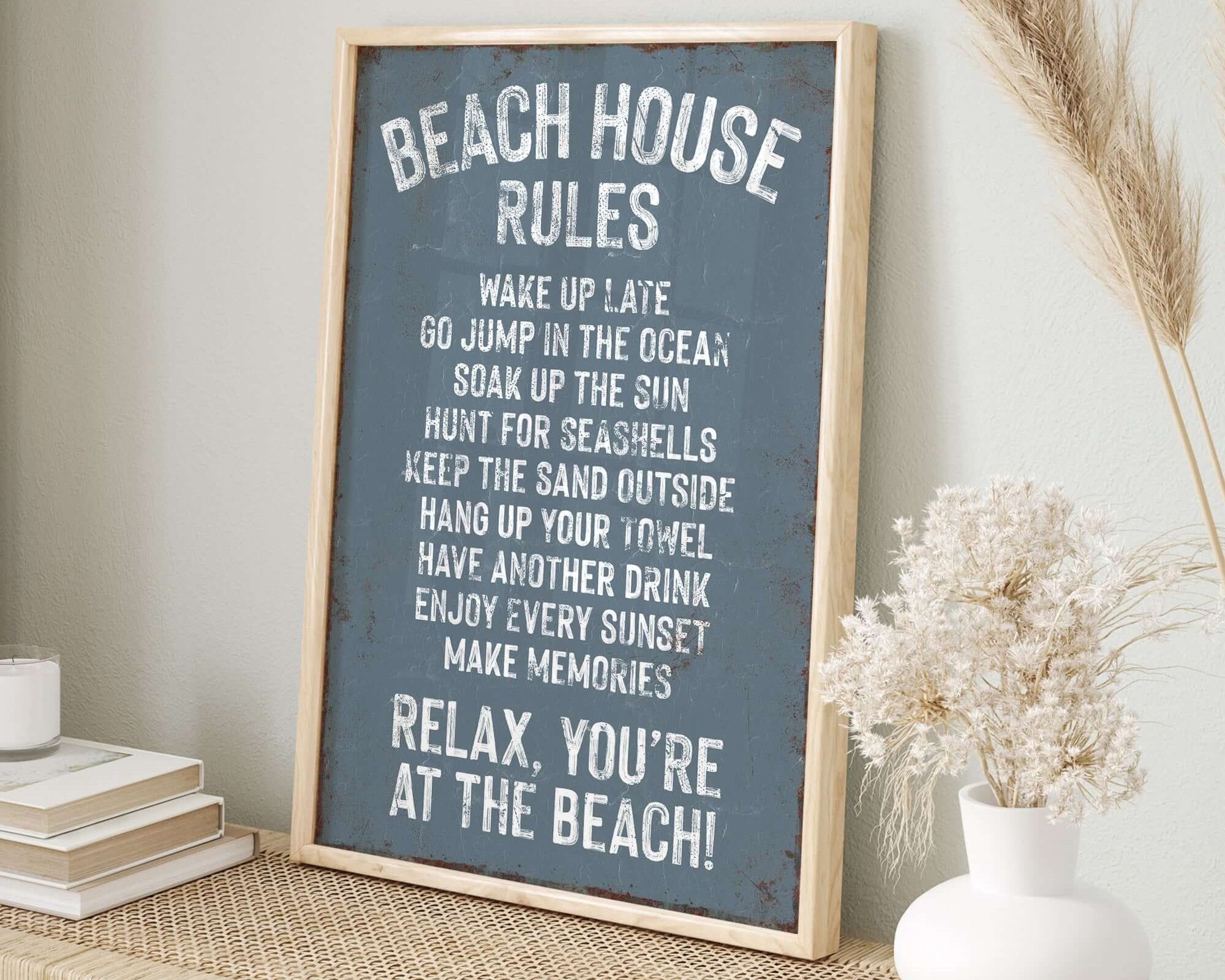 The "Beach House Rules Sign, Rusted Edge, Harbor Blue" brings vintage charm as wall art and home decor, beautifully displayed with beach accents on a wicker table against a light-colored wall.