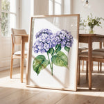 a framed painting of purple hydrangeas on a wooden table, with a vase of greenery and a wooden chair visible in the background.