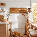A woman is standing in a kitchen, pointing to a framed sign that reads "Autumn Leaves and Apples Please" on the wall.