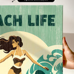 a hand holding a canvas print with a beach life theme, including a woman in a bathing suit and a wave.