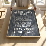 A person is holding up a sign with the text "BEACH HOUSE RULES" written on it, which appears to be a beach-themed sign with instructions for beachgoers.