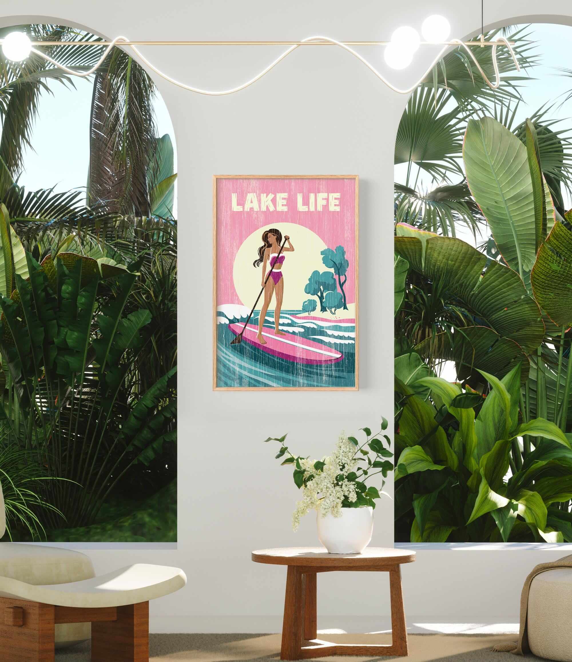 a poster of a woman paddleboarding on a tropical beach, with palm trees and a pink and yellow background. The poster is hanging on a wall, and there is a small table with a vase of flowers in front of it.