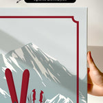 a hand holding a framed print of a mountain landscape with skis and ski poles.