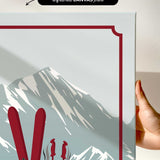a hand holding a framed print of a mountain landscape with skis and ski poles.