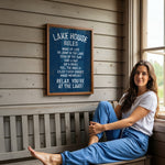 A woman is sitting on a bench in front of a sign that provides rules for the Lake House.