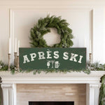 a fireplace mantel decorated with a wreath, candles, and a sign that reads "Apres Ski".