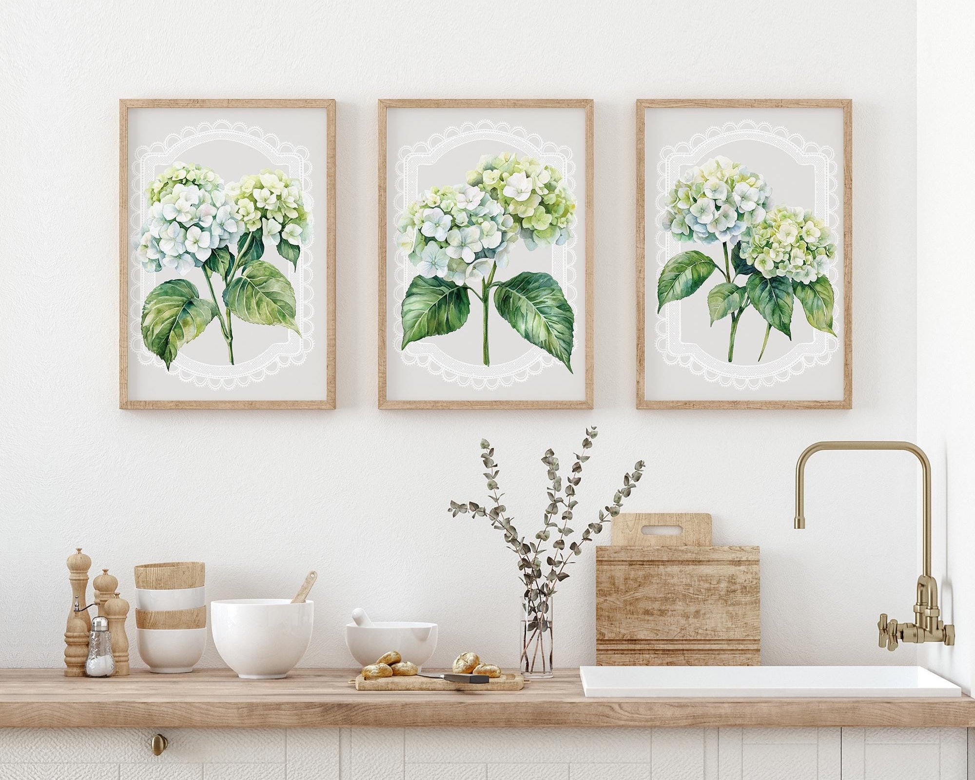 a kitchen counter with three framed watercolor paintings of hydrangeas hanging above it. The paintings are arranged in a row, creating a visually appealing display. On the counter, there are various kitchen items such as bowls, a vase with flowers, and a potted plant. The overall scene has a clean and organized appearance, with the framed paintings and kitchen items complementing each other.