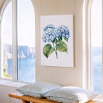 a serene indoor setting with a large window overlooking a picturesque view of the ocean. A painting of blue hydrangeas is displayed on the wall, adding a touch of nature to the space. In front of the window, there is a wooden bench with two pillows, providing a comfortable spot to sit and enjoy the view.