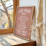 A framed sign on a window sill that reads "LAKESHOUSE RULES" and provides a list of activities to enjoy while at the lake, including "WAKE UP LATE", "GO JUMP IN THE LAST", "SINK IN THE SUN", "TAKE A NAP", "SIP A DRINK", "FEEL THE BREZE", "ENJOY EVERY SUNSET", and "MAKE MEMORIES".