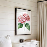 a framed painting of pink hydrangeas hanging on a white wall above a white dresser with a stack of books on top.