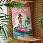 A framed poster with a woman in a pink bikini standing on a pink surfboard, holding a paddle, and surrounded by palm leaves.