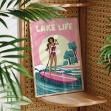 A framed poster with a woman in a pink bikini standing on a pink surfboard, holding a paddle, and surrounded by palm leaves.