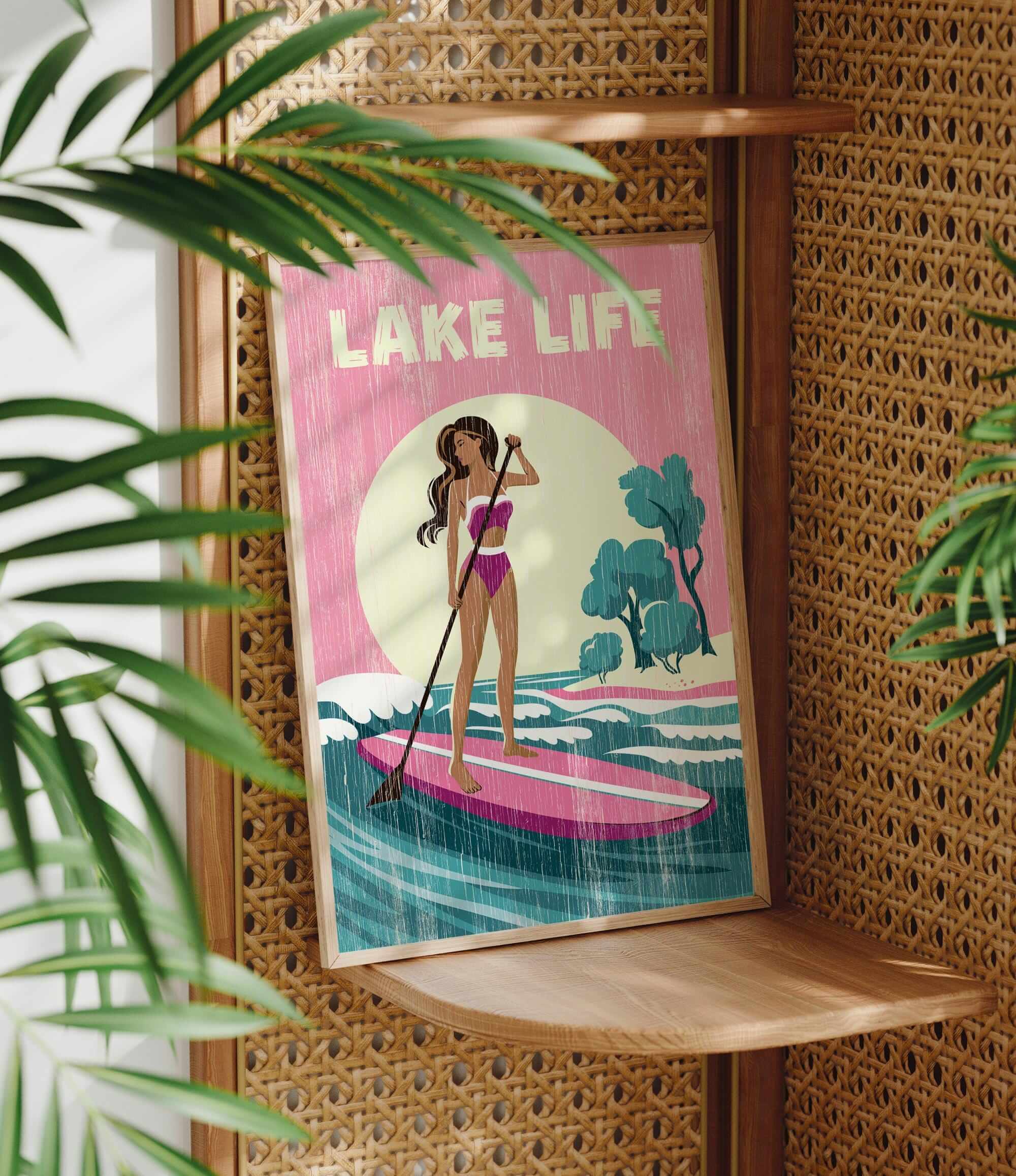 A framed poster with a woman in a pink bikini standing on a pink surfboard, holding a paddle, and surrounded by palm leaves.