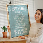 A woman is holding up a sign that says "LAKESHOUSE RULES" in front of a wooden table with a potted plant.