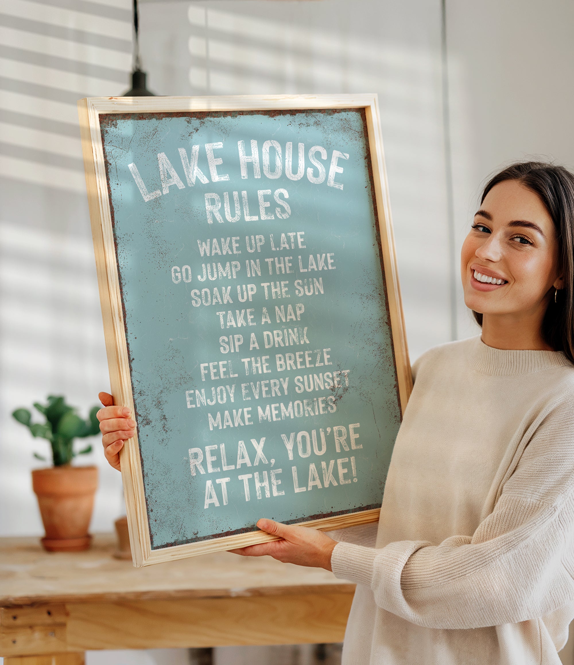 A woman is holding up a sign that says "LAKESHOUSE RULES" in front of a wooden table with a potted plant.