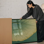 A woman in a gray sweater is leaning over a large framed picture, examining it closely.