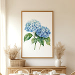 a dining room with a large framed painting of blue hydrangeas hanging on the wall above a wooden table. The table is set with white plates and cups, and there are two potted plants on either side of the table.