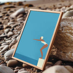 A framed illustration of a woman in a white swimsuit diving off a cliff, set against a blue background, is displayed on a rocky beach.
