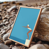 A framed illustration of a woman in a white swimsuit diving off a cliff, set against a blue background, is displayed on a rocky beach.