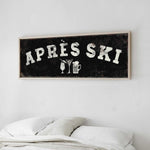 a framed sign with the text "APRES SKI" on it, hanging on a wall above a bed with white pillows.