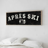a framed sign with the text "APRES SKI" on it, hanging on a wall above a bed with white pillows.