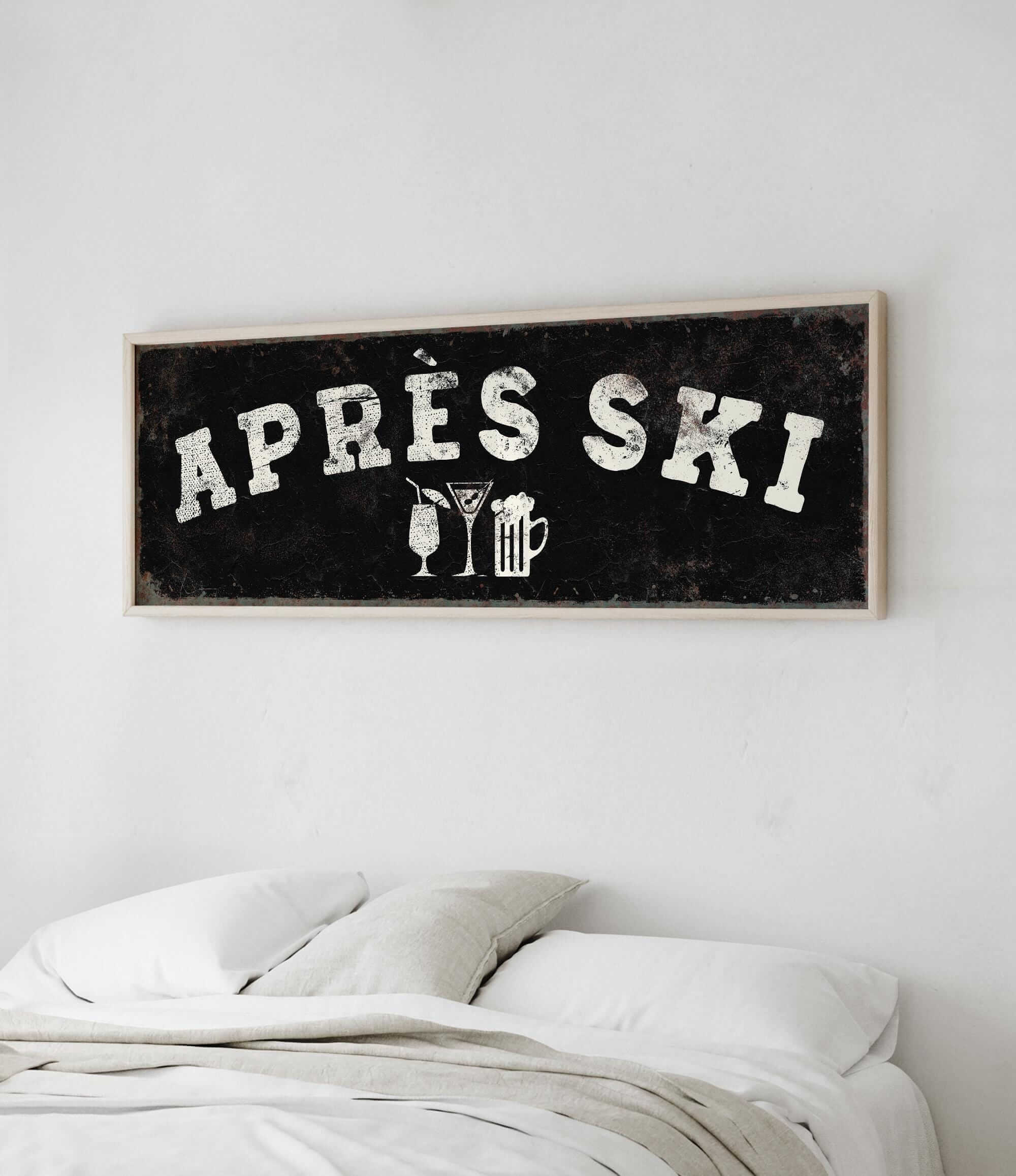 a framed sign with the text "APRES SKI" on it, hanging on a wall above a bed with white pillows.