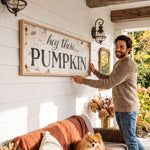A man is standing on a porch, holding up a large sign that says "hey there PUMPKIN" in front of a white wall. The porch has a couch, a dog, and some decorations, creating a cozy and festive atmosphere.