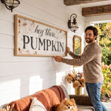 A man is standing on a porch, holding up a large sign that says "hey there PUMPKIN" in front of a white wall. The porch has a couch, a dog, and some decorations, creating a cozy and festive atmosphere.