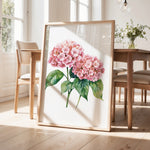 a framed painting of pink hydrangeas on a wooden table, with a vase of greenery and a wooden chair visible in the background.