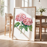 a framed painting of pink hydrangeas on a wooden table, with a vase of greenery and a wooden chair visible in the background.