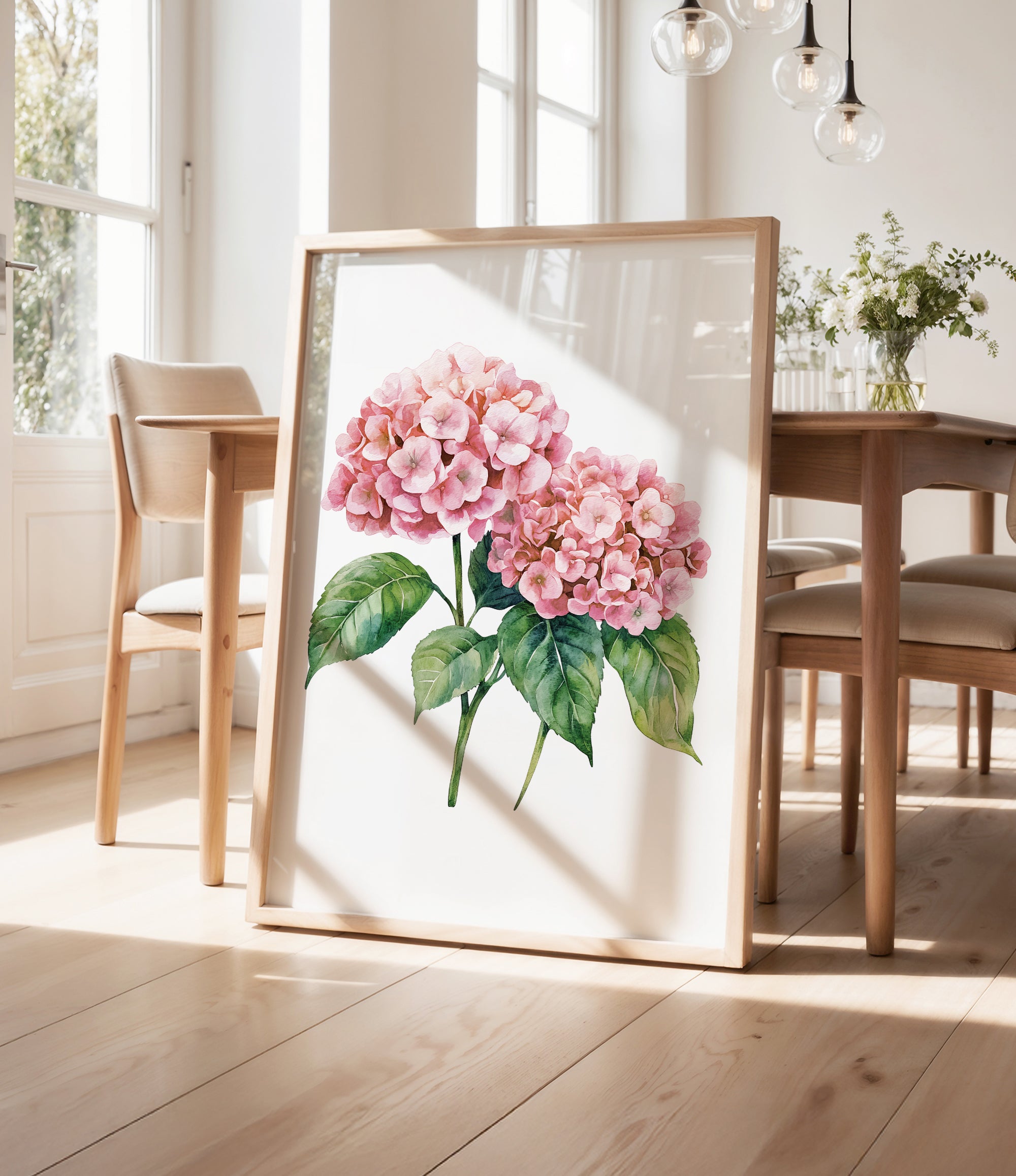 a framed painting of pink hydrangeas on a wooden table, with a vase of greenery and a wooden chair visible in the background.