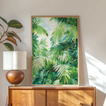 a wooden cabinet with a lamp on top, and a framed painting of lush green palm leaves hanging on the wall above the cabinet.