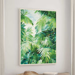 a large framed painting of lush green palm leaves hanging on a wall above a wooden table.