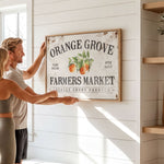 A man and a woman are hanging a vintage-style sign for "Orange Grove Farmers Market" on a white wall.