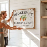 A man and a woman are hanging a vintage-style sign for "Orange Grove Farmers Market" on a white wall.