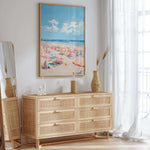 a beach scene with a large framed painting hanging on the wall above a wooden dresser with six drawers. The dresser has a wicker vase and a bottle on top, and there is a mirror on the left side of the dresser. The room has a bright and airy atmosphere, with natural light coming in through the window and a white curtain.