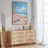 a beach scene with a large framed painting hanging on the wall above a wooden dresser with six drawers. The dresser has a wicker vase and a bottle on top, and there is a mirror on the left side of the dresser. The room has a bright and airy atmosphere, with natural light coming in through the window and a white curtain.