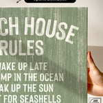A hand holds a Beach House Rules Sign, crafted from painted wood in seagrass color with white text, perfect as beach house wall decor and bathed in sunlight.