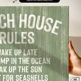 A hand holds a Beach House Rules Sign, crafted from painted wood in seagrass color with white text, perfect as beach house wall decor and bathed in sunlight.