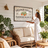A woman in a white dress is standing in a room with a large framed sign that reads "Orange Grove Farmers Market" and various potted plants surrounding the space.