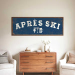 a living room with a large blue sign that says "APRÈS SKI" above a wooden dresser. The dresser has a vase of flowers and a book on it. There are two white chairs in the room, and a small table with a lamp on it.
