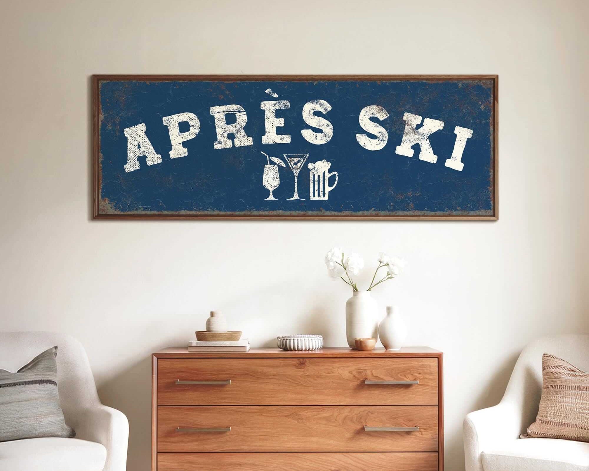 a living room with a large blue sign that says "APRÈS SKI" above a wooden dresser. The dresser has a vase of flowers and a book on it. There are two white chairs in the room, and a small table with a lamp on it.