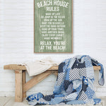 A painted wood Beach House Rules Sign with seagrass hangs above a bench with a blue patchwork quilt, bringing charming coastal wall art to the bright room.