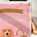 a hand holding a framed canvas print with a dog illustration on it. The text on the canvas print reads "Don't forget to wash your paws".