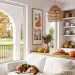 A cozy living room with a white sofa, a dog sleeping on the armrest, and a wicker pendant light hanging above. The room has a window overlooking a golf course, and there are various fall-themed decorations and items, such as a framed sign, a vase of flowers, and a bowl of fruit.
