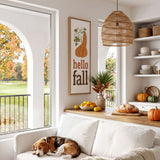 A cozy living room with a white sofa, a dog sleeping on the armrest, and a wicker pendant light hanging above. The room has a window overlooking a golf course, and there are various fall-themed decorations and items, such as a framed sign, a vase of flowers, and a bowl of fruit.