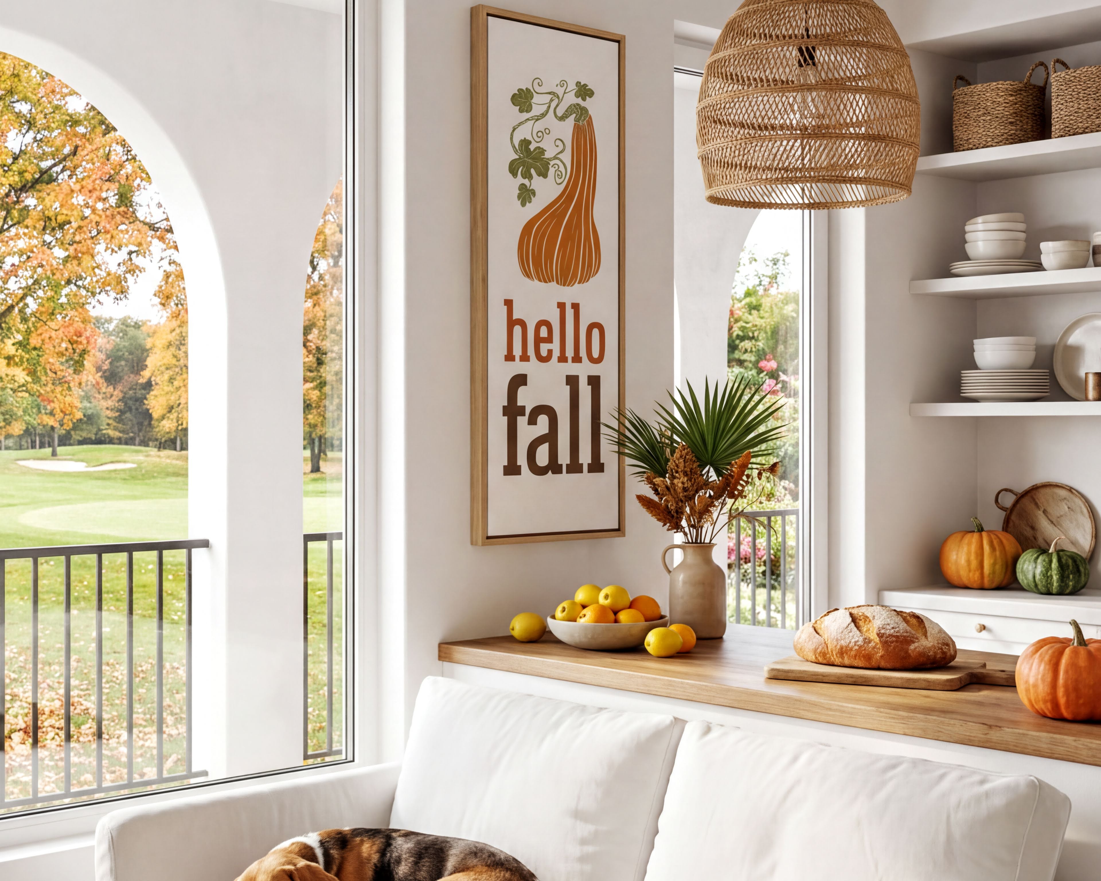 A cozy living room with a white sofa, a dog sleeping on the armrest, and a wicker pendant light hanging above. The room has a window overlooking a golf course, and there are various fall-themed decorations and items, such as a framed sign, a vase of flowers, and a bowl of fruit.