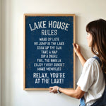A woman is hanging a framed sign on a wall that lists the "Lake House Rules" for a relaxing lakeside experience.