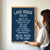 A woman is hanging a framed sign on a wall that lists the "Lake House Rules" for a relaxing lakeside experience.
